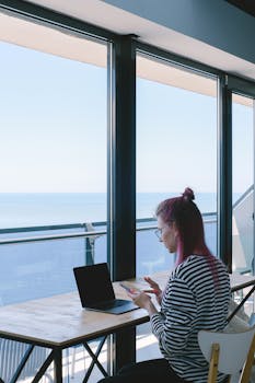 Woman with pink hair working on laptop by ocean view window in modern setting.