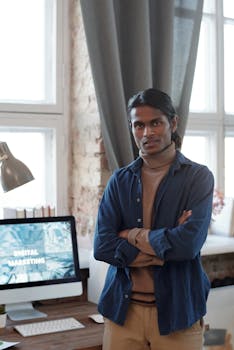 Young man posing confidently in a contemporary home office setting with digital marketing displayed on the computer screen.