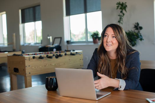 A businesswoman in a black blazer working on her laptop in a modern office setting.